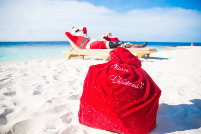 Santa Claus relaxing on a beach lounger at a tropical resort, with a red 'Merry Christmas' sack in the sand.