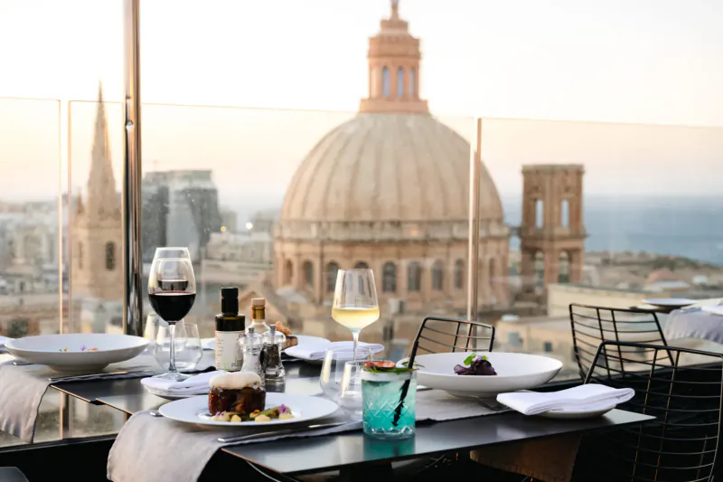 Rooftop table at Embassy Valletta Hotel with wine, cake, cocktail overlooking Valletta's domes, spires, and sea at dusk