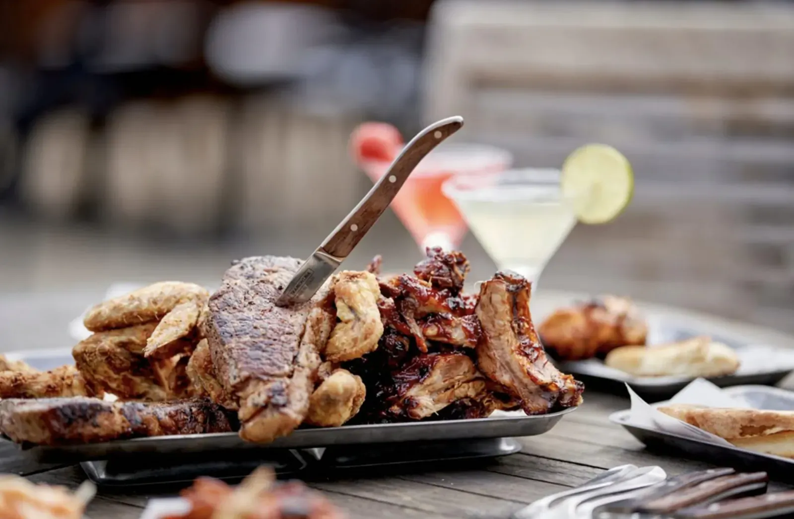 Platter of grilled ribs, steak, and chicken on wooden table with knife, margarita, and lime garnish