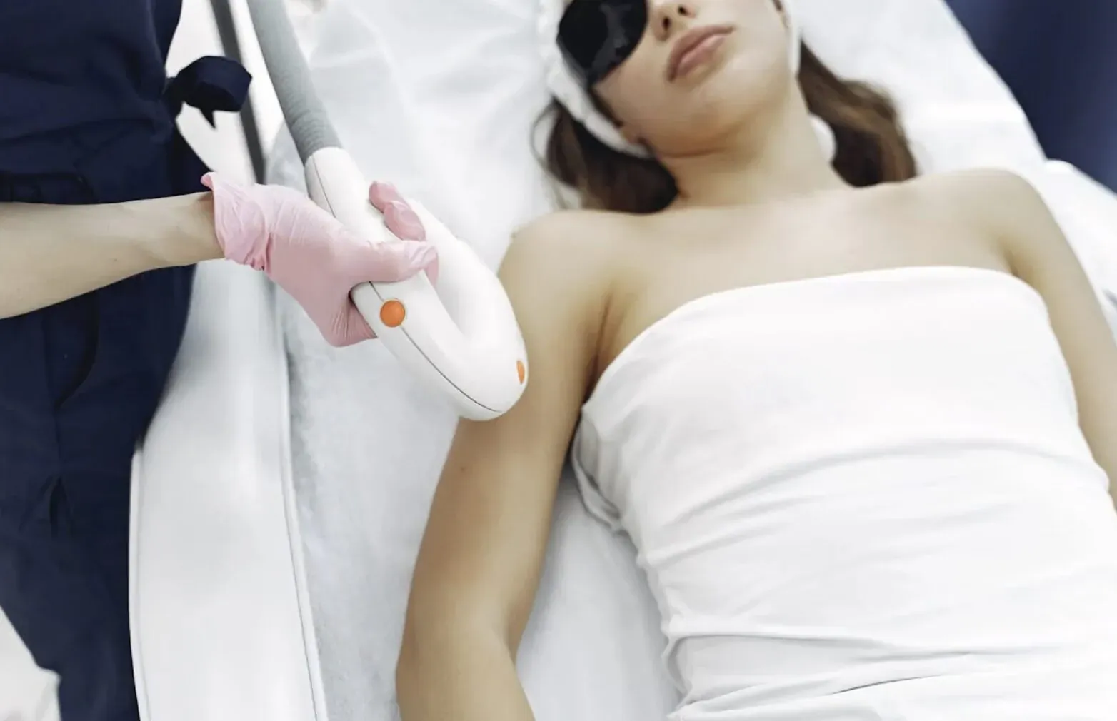 Laser technician in gloves applies laser hair removal device to woman's arm during session, patient wears eye protection on bed.