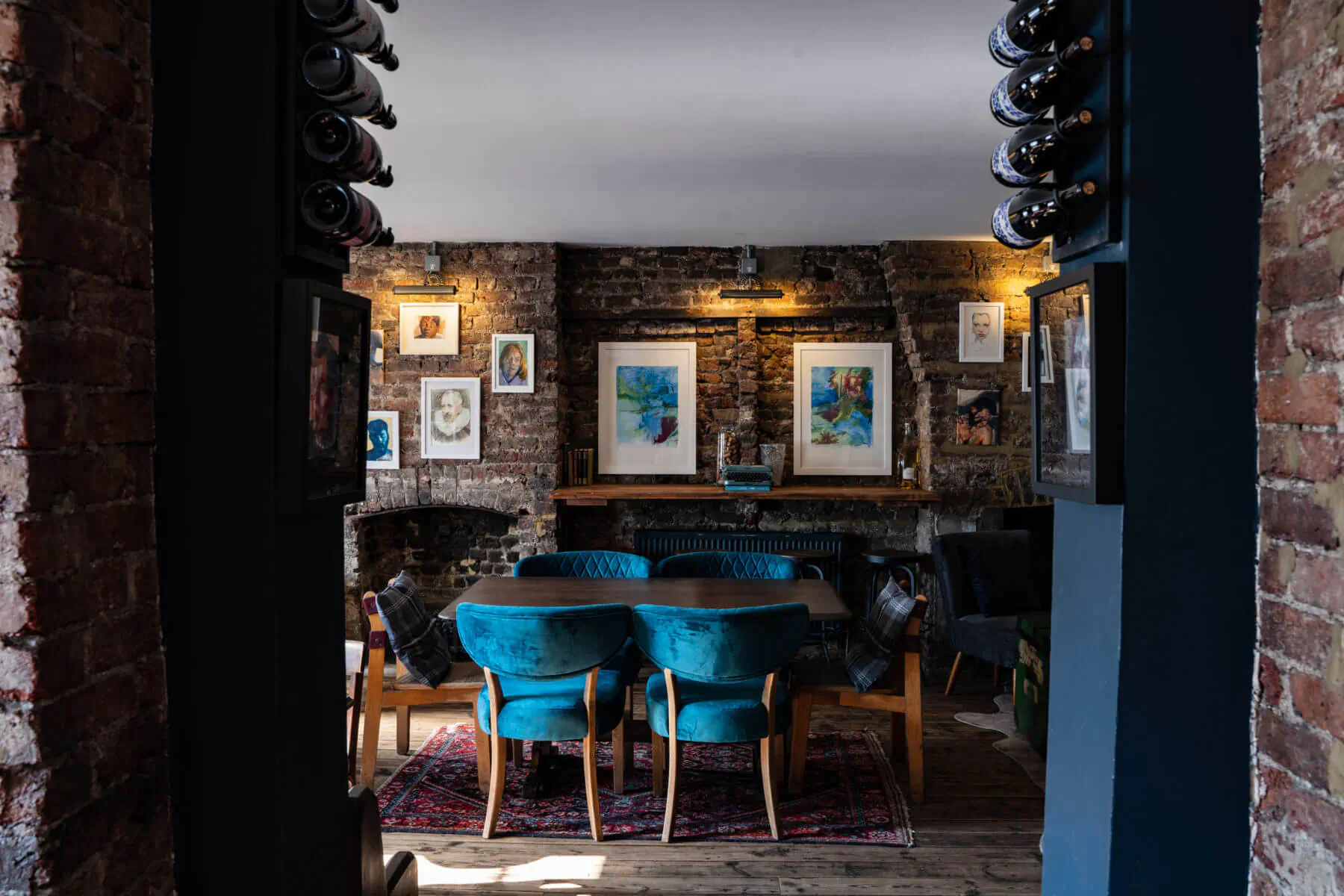 Cozy brick-walled dining room with teal velvet chairs around wooden table, wine racks, art, and warm lighting.