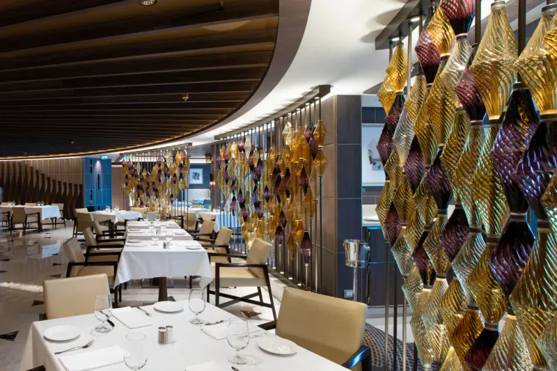 Elegant Princess Cruises dining room with white-clothed tables, gold and purple glass wall sculptures, curved ceiling.