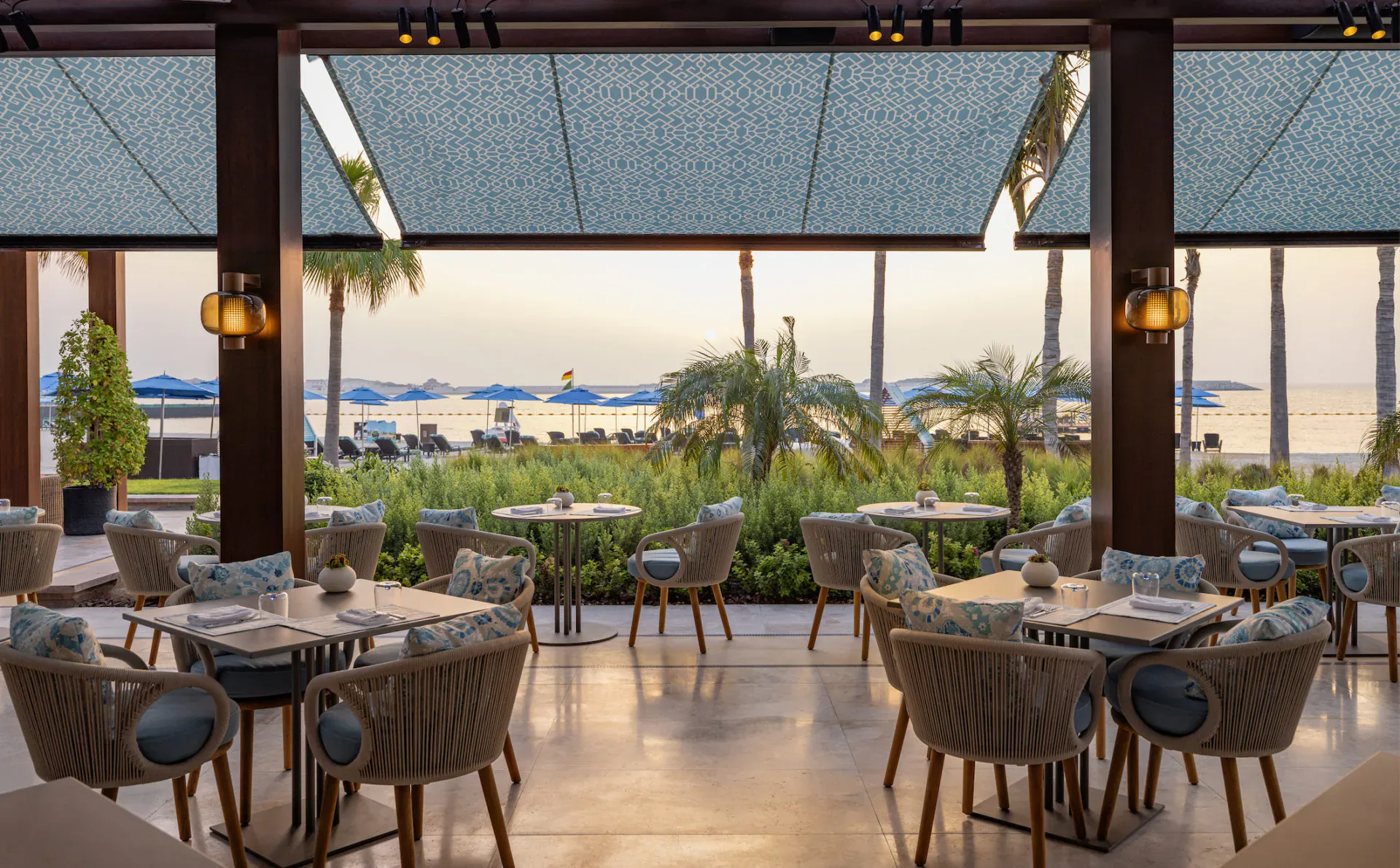 Open-air Sea Fu restaurant at Four Seasons Resort Dubai: wicker tables with blue cushions, lush garden, beach, palms at sunset.