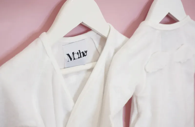 Two white baby onesies on hangers against pink wall, labeled 'MOTHER', one with bow detail.