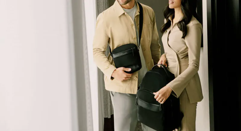 Smiling man in beige shirt and woman in beige suit holding black Antler Fenchurch bags by door