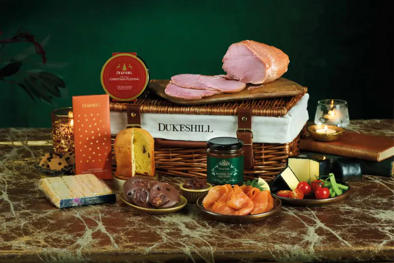 DukesHill wicker hamper on green table with sliced ham, cheese, salmon, chutney, biscuits, and candles.