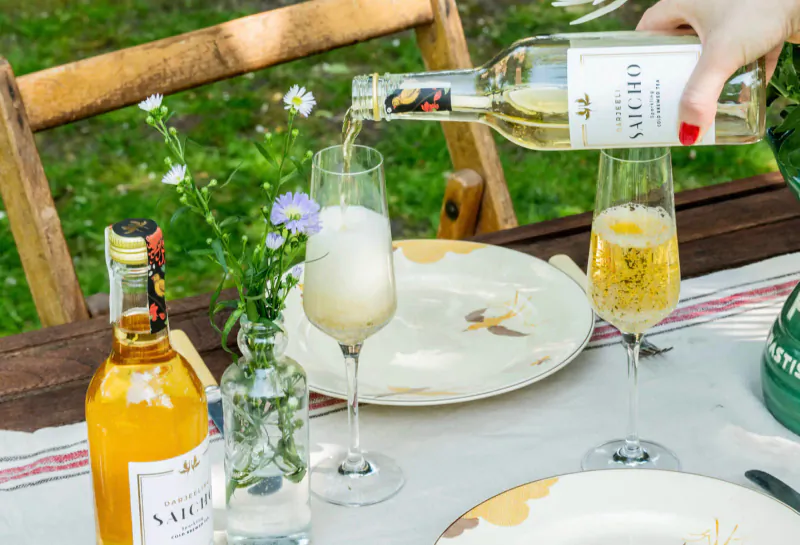 Hand pouring Saicho sparkling white tea from bottle into flute glass on outdoor wooden table with yellow tea bottle and plates