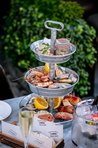 Three-tier metal tray with shrimp, mussels, oysters, lemon slices, and garnishes on outdoor table with champagne and 'Lobster Pot Booth' sign at The Mayfair Chippy