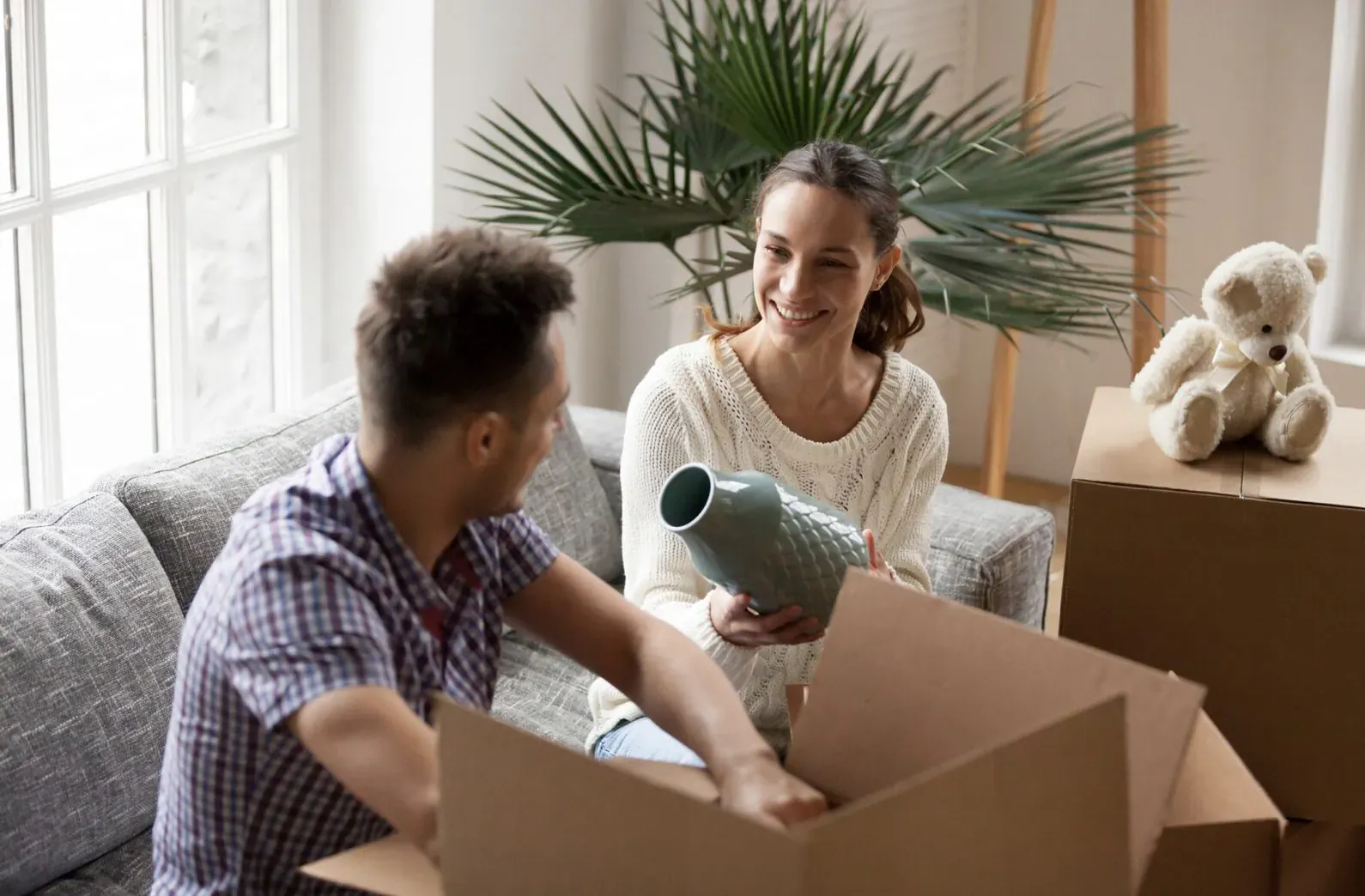 Smiling couple unpacking vase from box and teddy bear in new living room with plants and window.