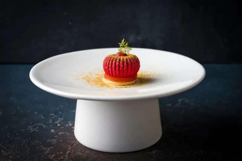 Red tomato-shaped dessert topped with herb sprig and gold dust on white pedestal plate