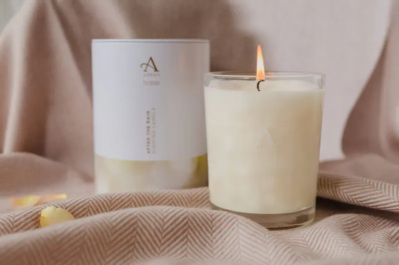 Lit white ARRAN Sense of Scotland candle in glass jar beside its white and gold box on beige fabric with yellow petals