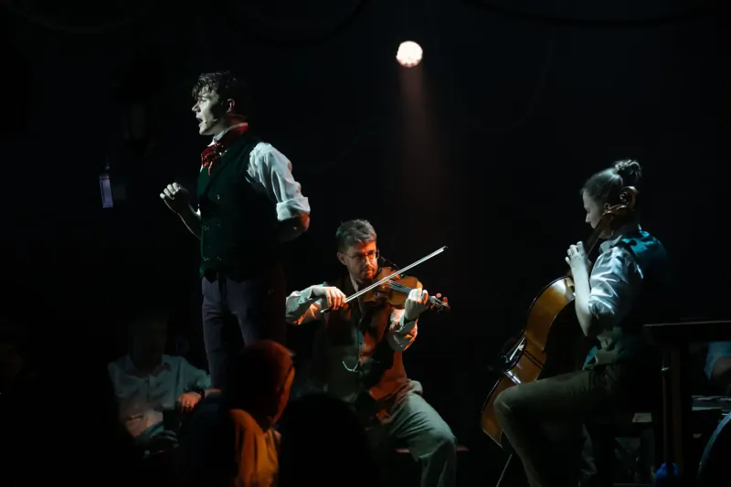 Performers in Victorian attire playing violin and cello on dimly lit stage at immersive Christmas dining event