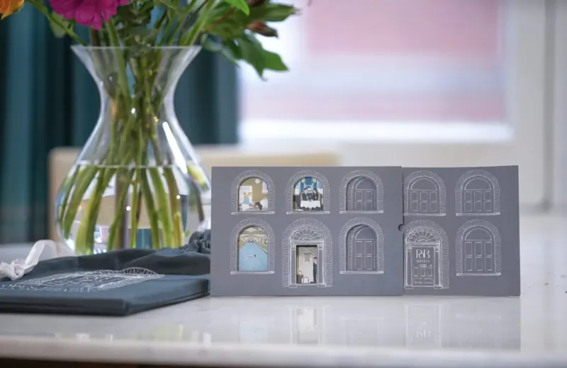 Gray 3D house facade with window scenes on table beside pink flowers in glass vase and blue cloth, soft pink background.