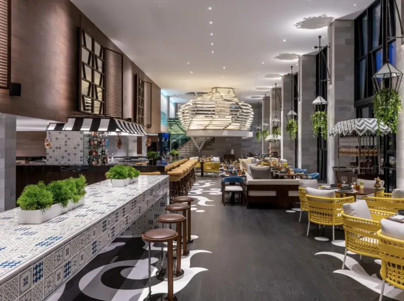 Spacious modern hotel restaurant at W Algarve with white marble bar, yellow chairs, plants, and large hanging light fixture.