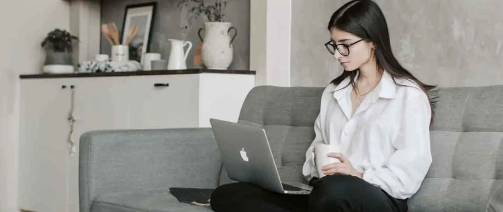 Young woman in white blouse working on MacBook laptop with coffee mug on gray sofa in modern kitchen