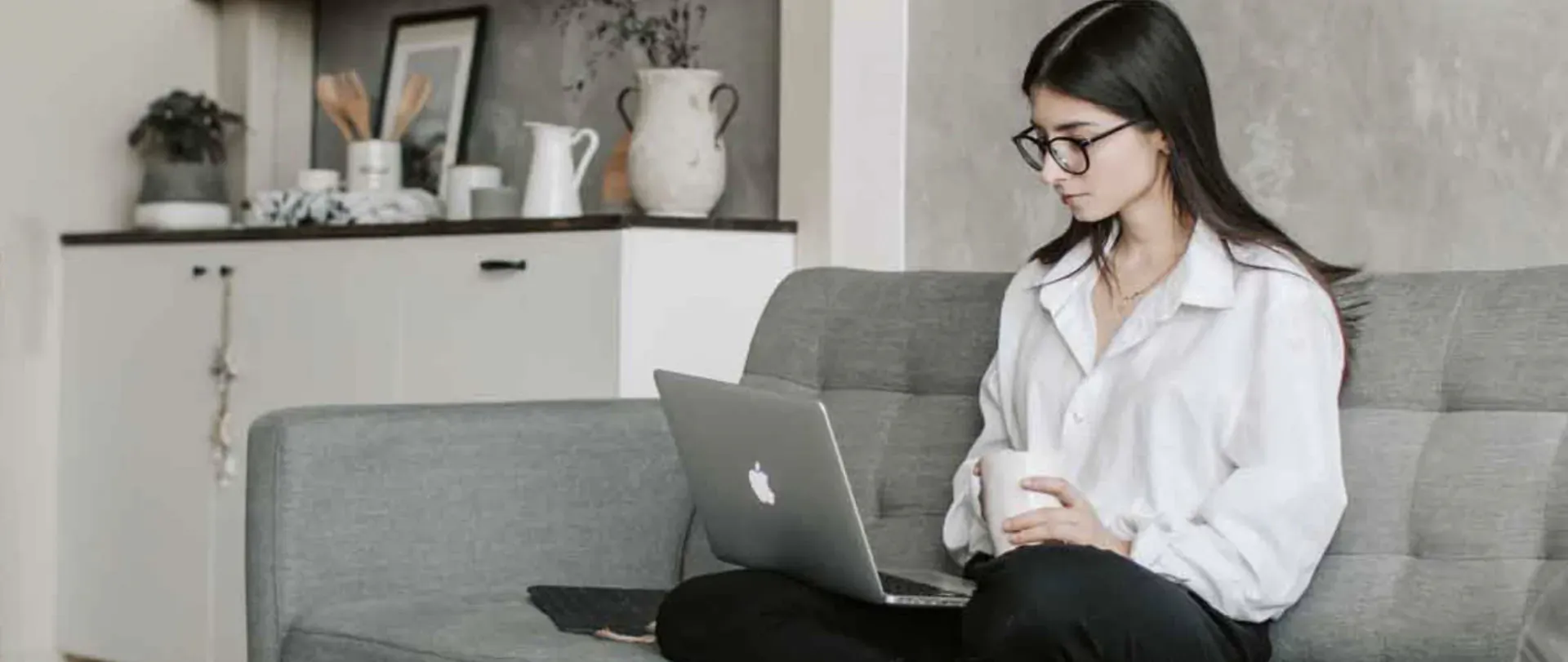 Young woman in white blouse working on MacBook laptop with coffee mug on gray sofa in modern kitchen