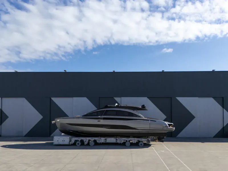Silver Pershing GTX70 yacht on trailer in front of modern gray warehouse with white chevron doors, blue sky.