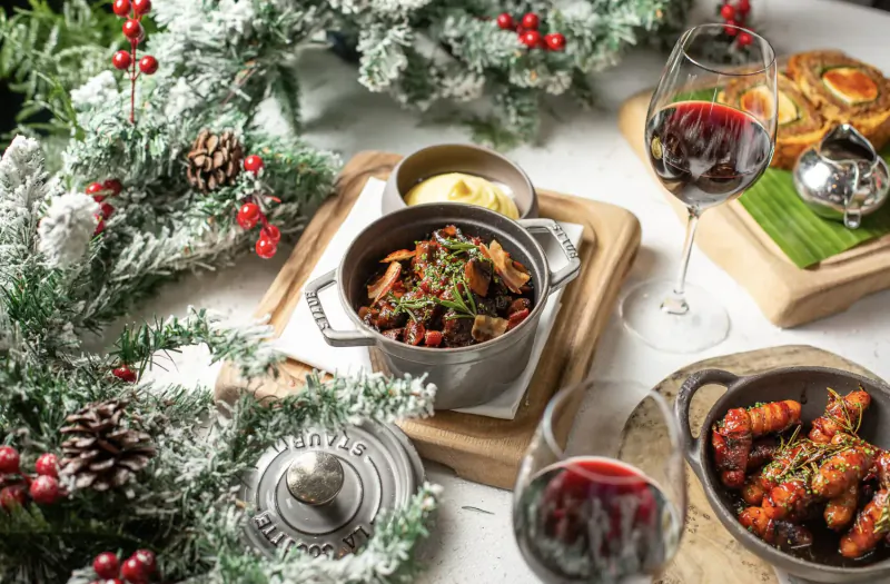 Festive Christmas table with braised meat stew, prawns, bread rolls, butter, and red wine amid greenery and berries.