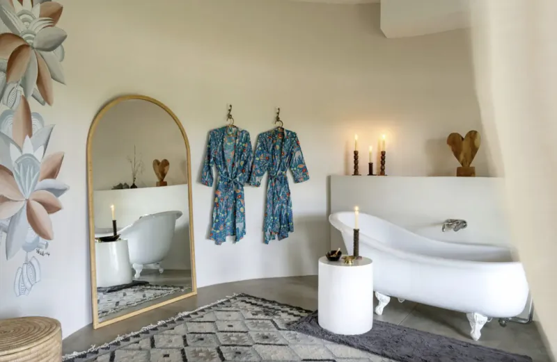 Elegant bathroom with clawfoot tub, gold arched mirror, blue floral robes on hooks, candles, and floral wall art.