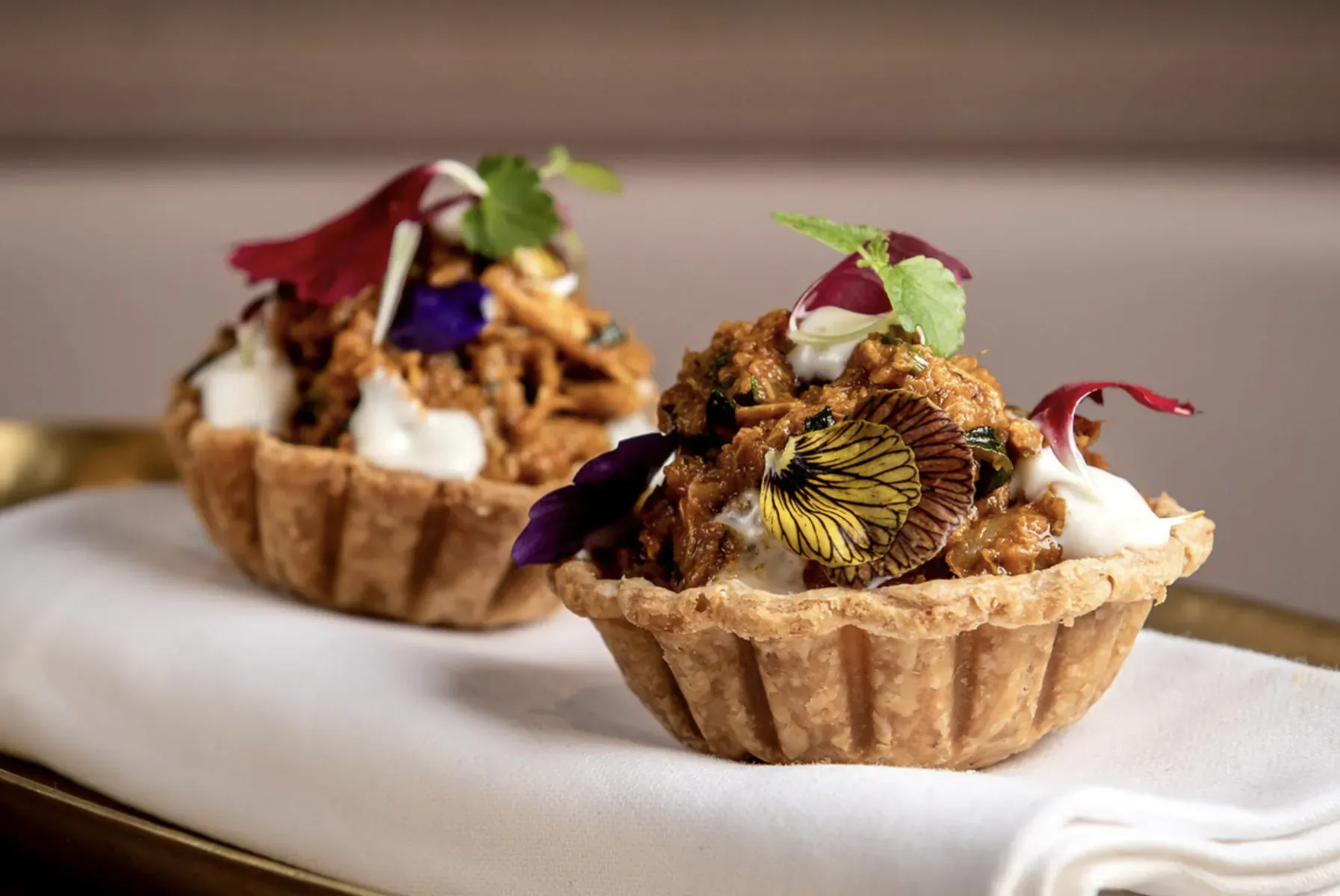 Two golden pastry tartlets filled with curried shredded meat, topped with cream, edible flowers, and herbs on a white napkin.