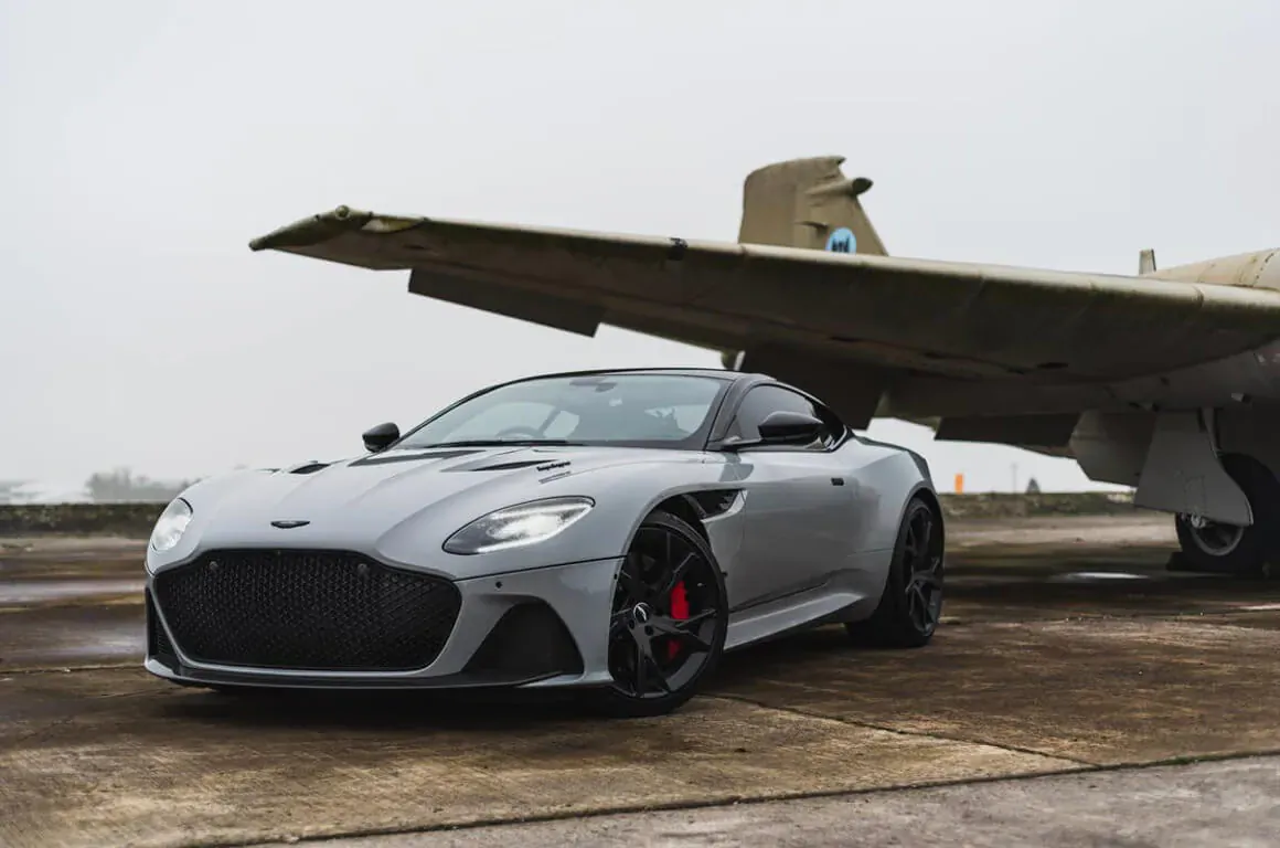 Silver Aston Martin sports car parked on tarmac under gray military airplane wing, foggy day.
