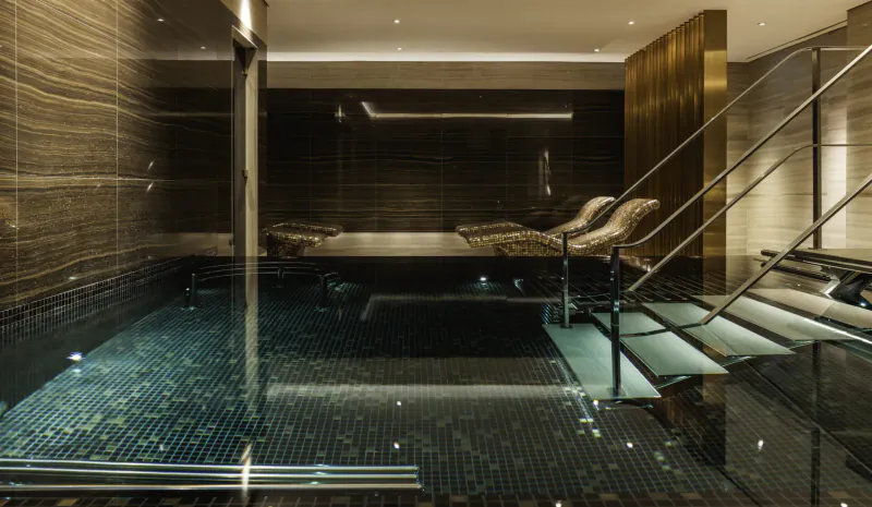 Luxurious indoor pool at The Langley hotel with mosaic tiles, lounge chairs, gold accents, and warm lighting.