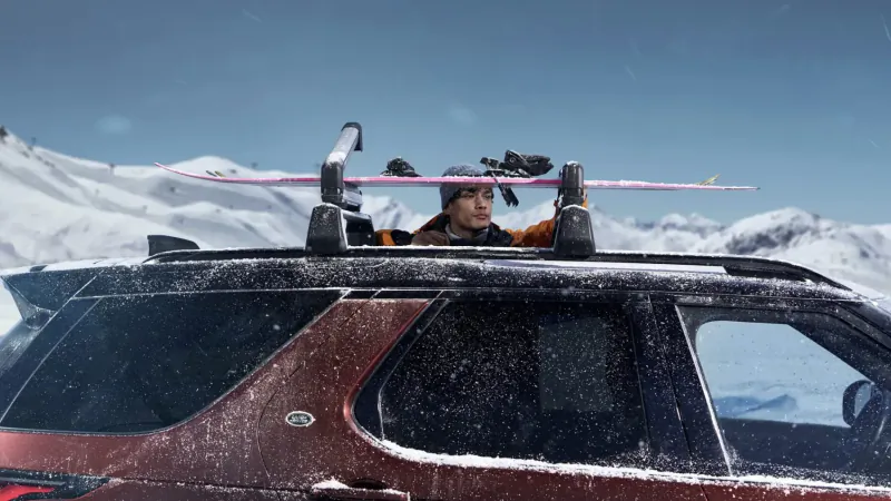 Woman in orange jacket peers from red JLR Discovery SUV roof rack with pink skis, snowy mountains backdrop