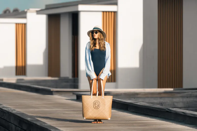 Woman in blue swimsuit and open shirt stands on wooden pier at Maldives resort, holding large tan tote bag, wearing sun hat