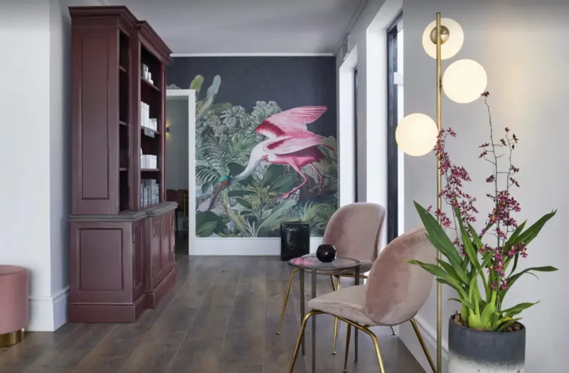 Luxurious hotel hallway with dark wood bookshelf, pink bird mural, velvet chairs, orchids, and floor lamp.