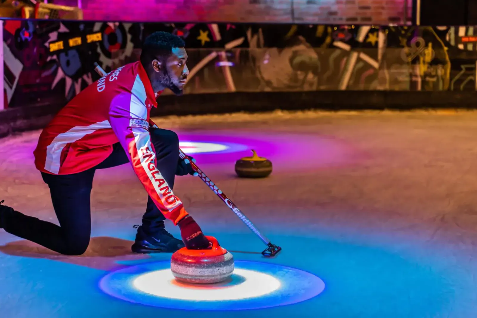 Player in red 'Queens' jacket crouches delivering curling stone on icy rink with purple lights, Beijing Winter Olympics event in London