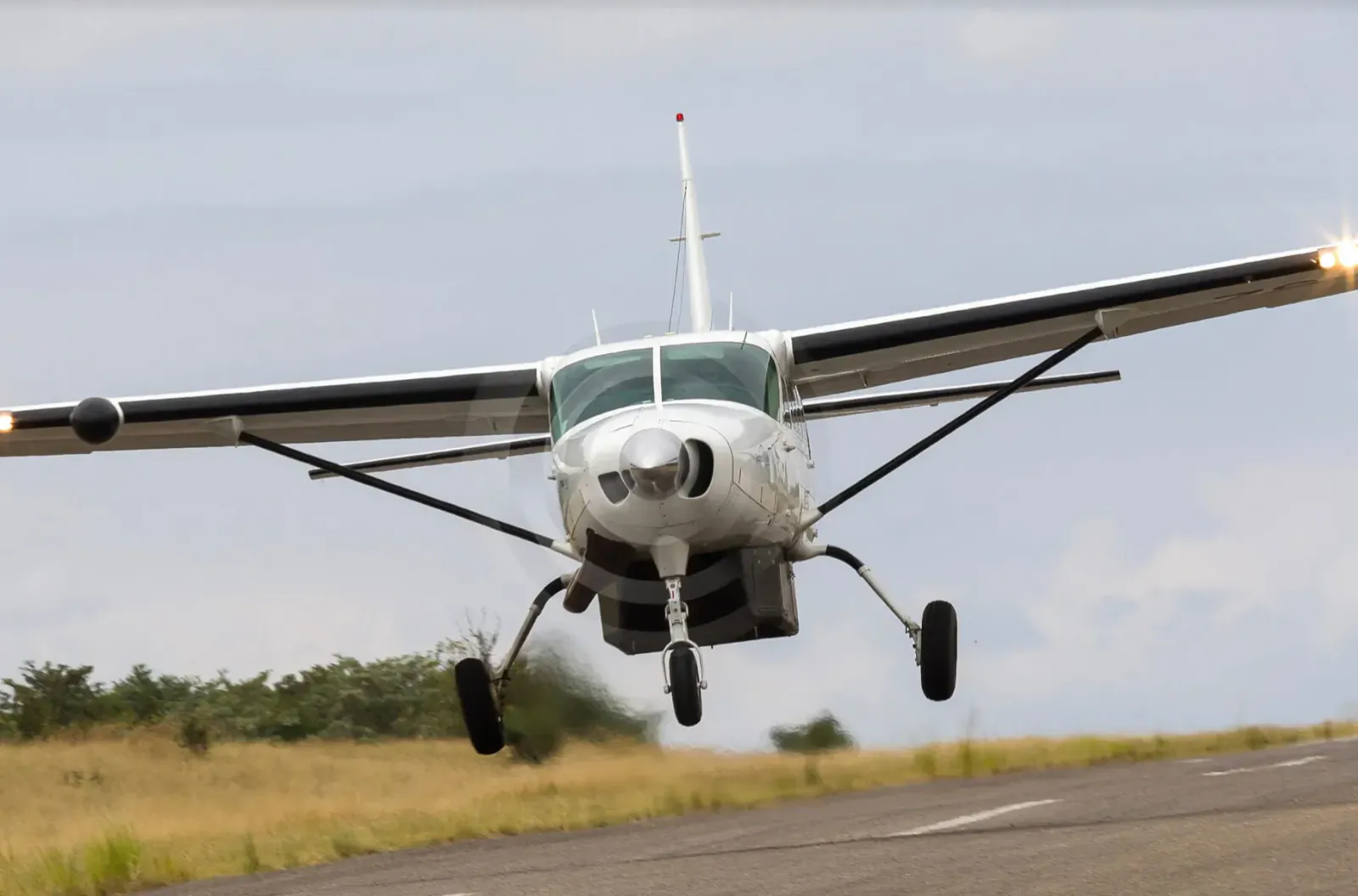 White Cessna airplane taking off on runway with landing lights on, grassy field backdrop