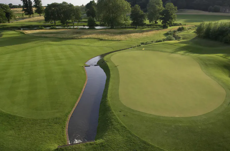Aerial view of lush golf course with winding stream between two manicured greens and bunkers amid trees.