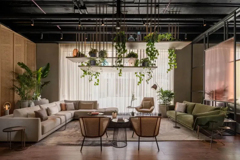 Modern loft lounge with beige L-shaped sofa, green armchair, hanging plants, wood floors, and large windows.