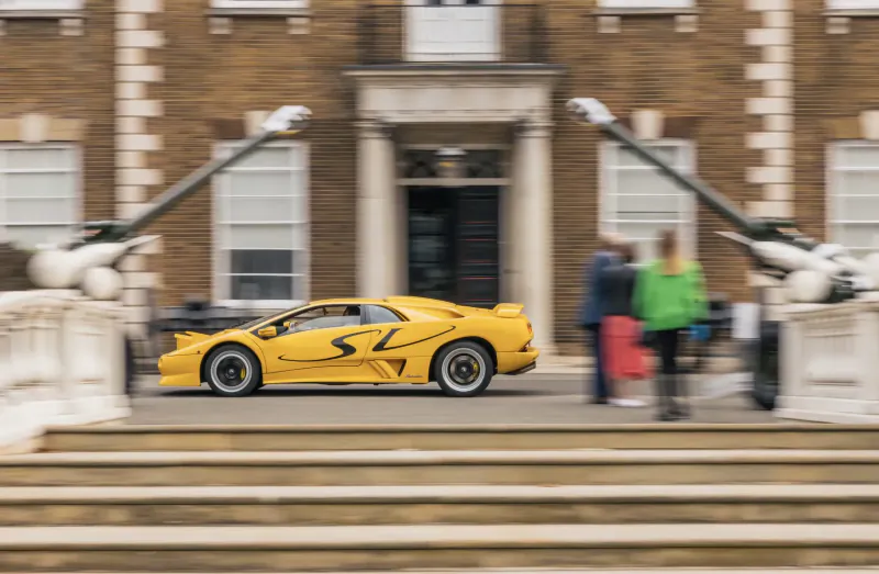 Yellow Lamborghini Diablo VT with S racing stripes parked before grand brick mansion at London Concours 2024, people nearby.
