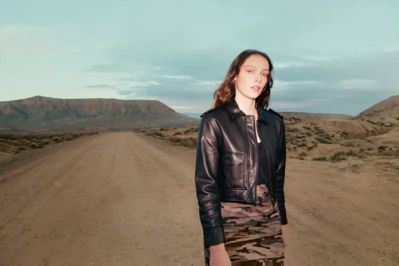 Woman in black leather jacket and camo skirt stands on desert road amid mountains at dusk
