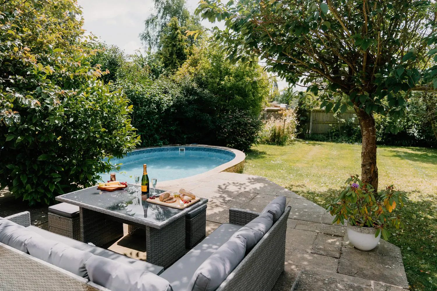 Outdoor patio at Cotswolds country house with gray sofa, charcuterie board, champagne by kidney-shaped pool and lush garden.