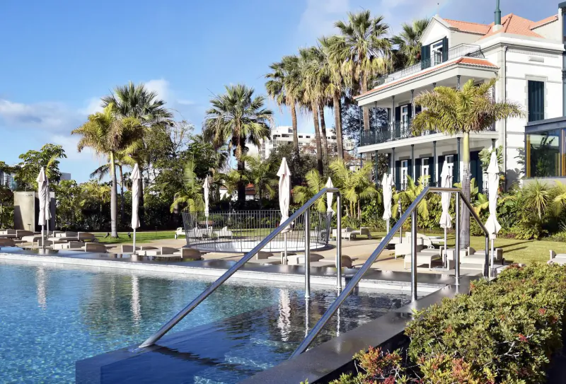 Infinity pool with stairs at Les Suites Hotel at The Cliff Bay, Funchal, Madeira, surrounded by palms and white colonial building under blue sky