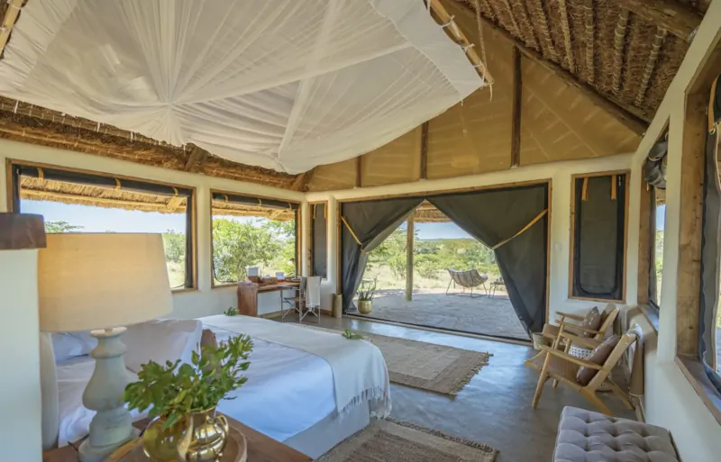 Luxurious safari tent interior with white canopy bed, wooden accents, chairs, desk, and savanna view through large windows.