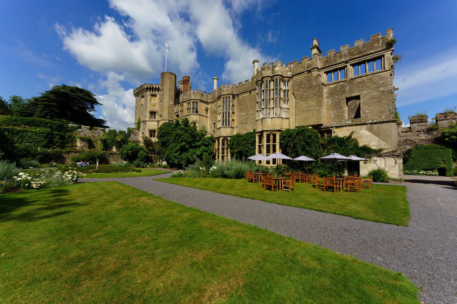 Thornbury Castle hotel in Bristol, grand stone facade with turrets, gardens, and outdoor cafe tables under blue sky.