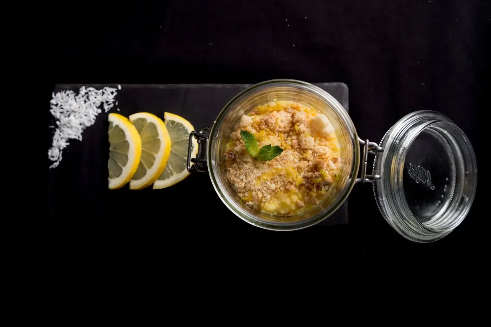Glass jar of yellow creamy dessert topped with granules and mint, garnished with lemon slices on salt-edged black slate