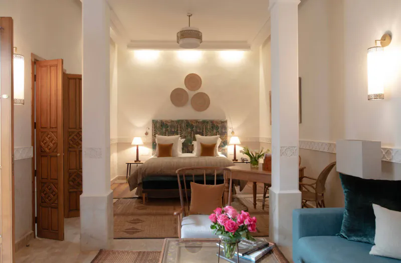 Elegant Marrakech hotel suite with tufted bed, velvet sofa, pink roses, woven wall art, and arched doorways.