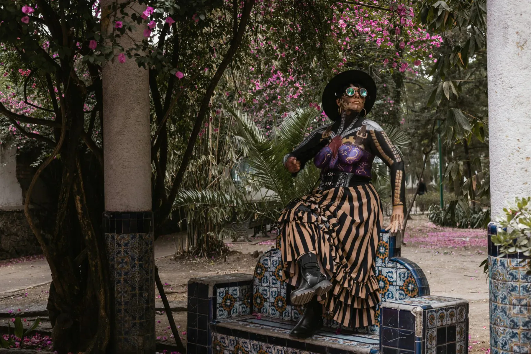 Colorful skeleton woman statue in wide hat and striped skirt, sitting on tiled bench amid pink bougainvillea and trees