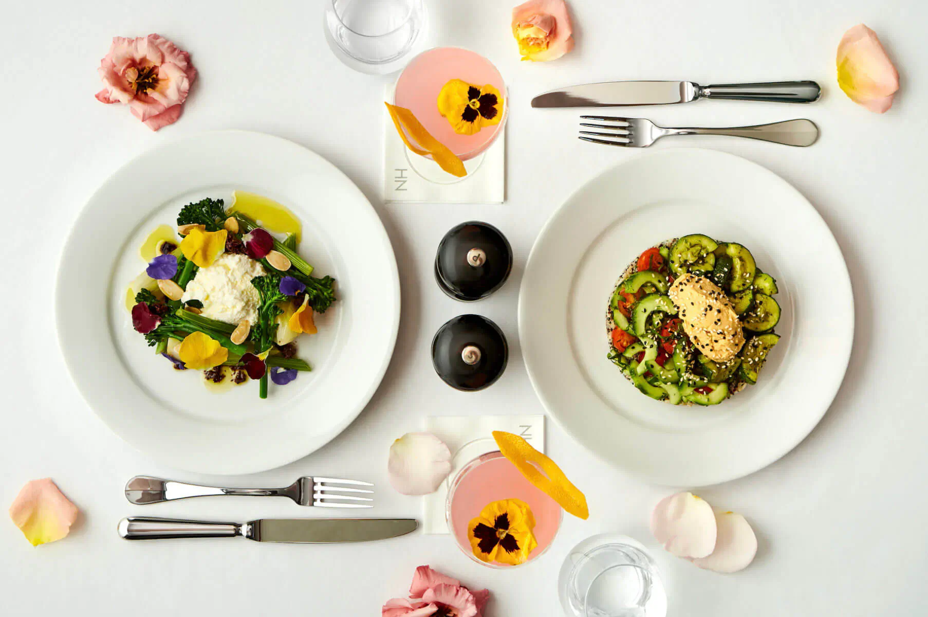 Top-down view of two plates: colorful veggie salad with feta and zucchini pasta with cream, flanked by pink cocktails, cutlery, pepper mills, and rose petals.