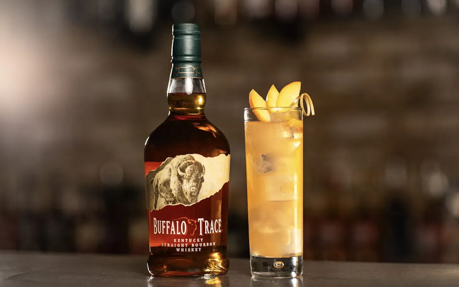 Buffalo Trace bourbon bottle with buffalo label beside tall glass of iced drink garnished with orange slices on bar