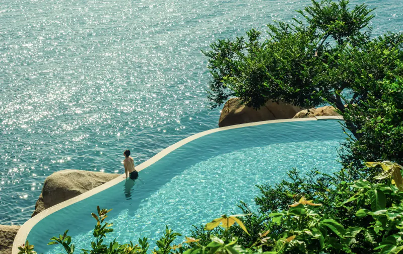 Person stands at edge of curved infinity pool overlooking turquoise sea and lush greenery at Silavadee Pool Spa Resort
