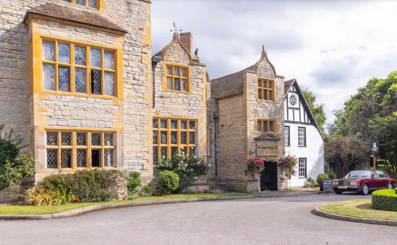 Karma Salford Hall, Tudor-style stone manor with half-timbered wing, flowers, cars on driveway in Vale of Evesham.