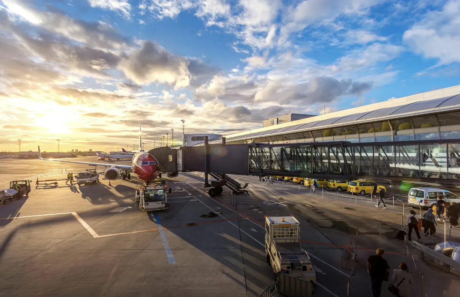 Jet bridge connected to red airplane at modern airport terminal during golden hour sunset with ground crew.