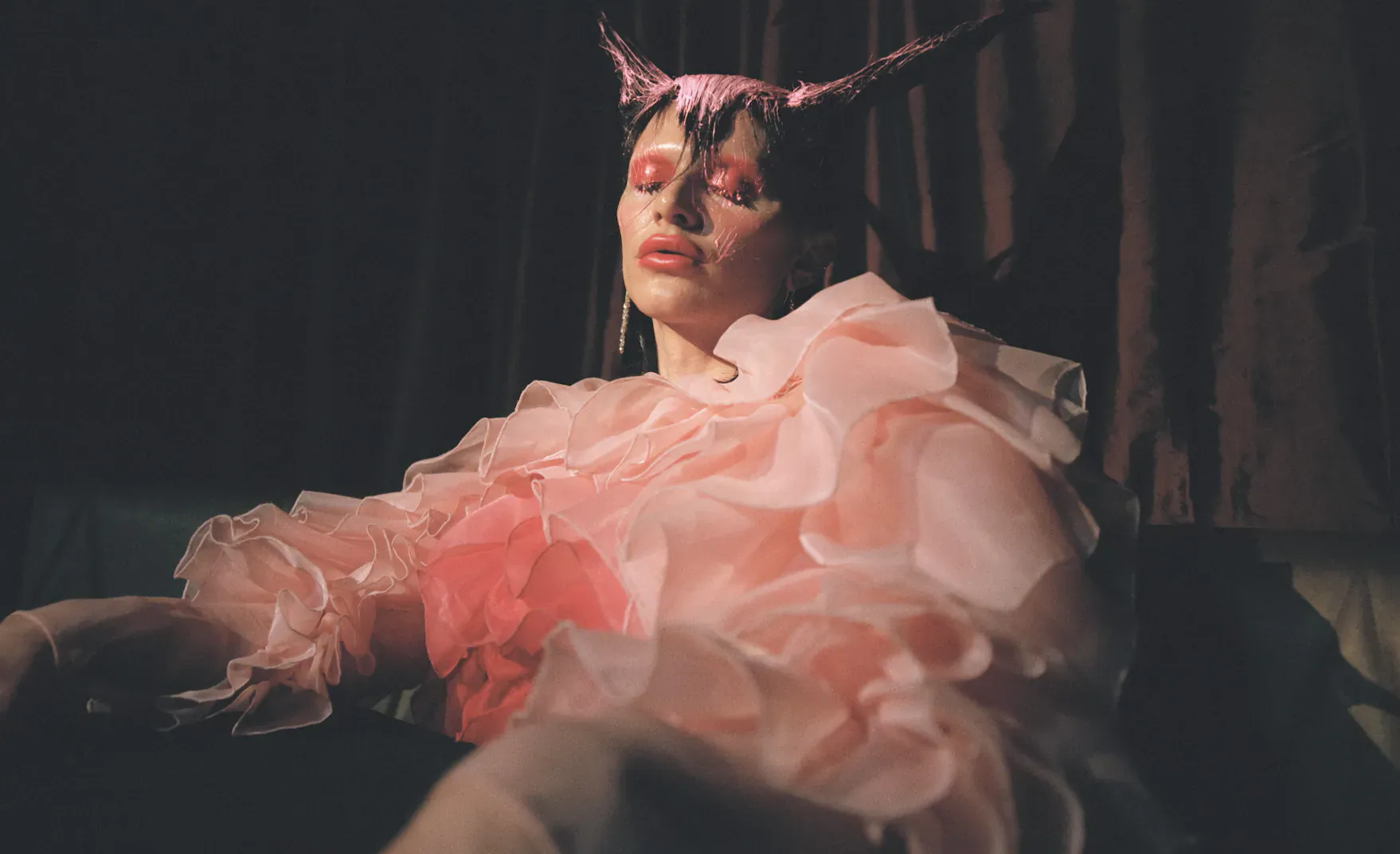 Woman with pink demon horns and bold makeup reclines in ruffled pink gown on sofa in dim room