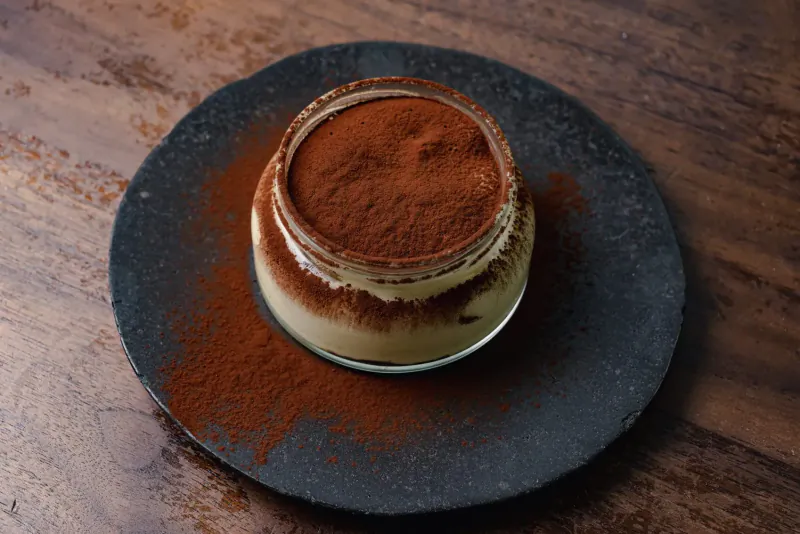 Tiramisu in glass jar topped with cocoa powder, on black plate over wooden table