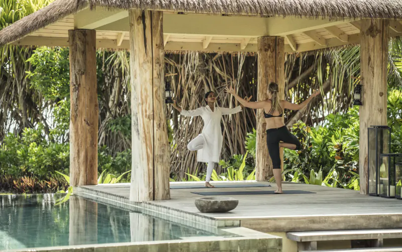 Man in white tunic and woman in black leggings practicing yoga poses on a pavilion deck by infinity pool amid tropical palms at Four Seasons resort.